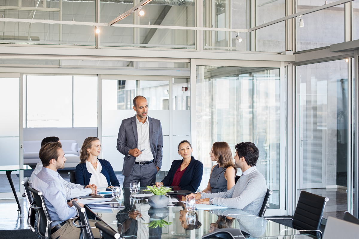 group of business people sitting around a glass table with one man standing